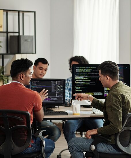 Group of people collaborating at a table with computer screens displaying code in a modern office setting.